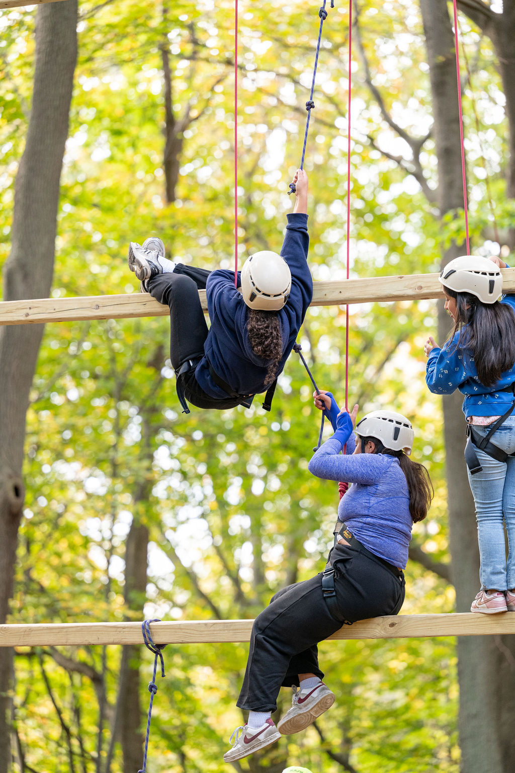 The image shows three people on a ropes course in a forest. One person is hanging from a rope, while another is climbing. A third person is standing on a platform. They are all wearing helmets and harnesses for safety. The course is made of wooden beams and ropes, and the background is filled with trees.